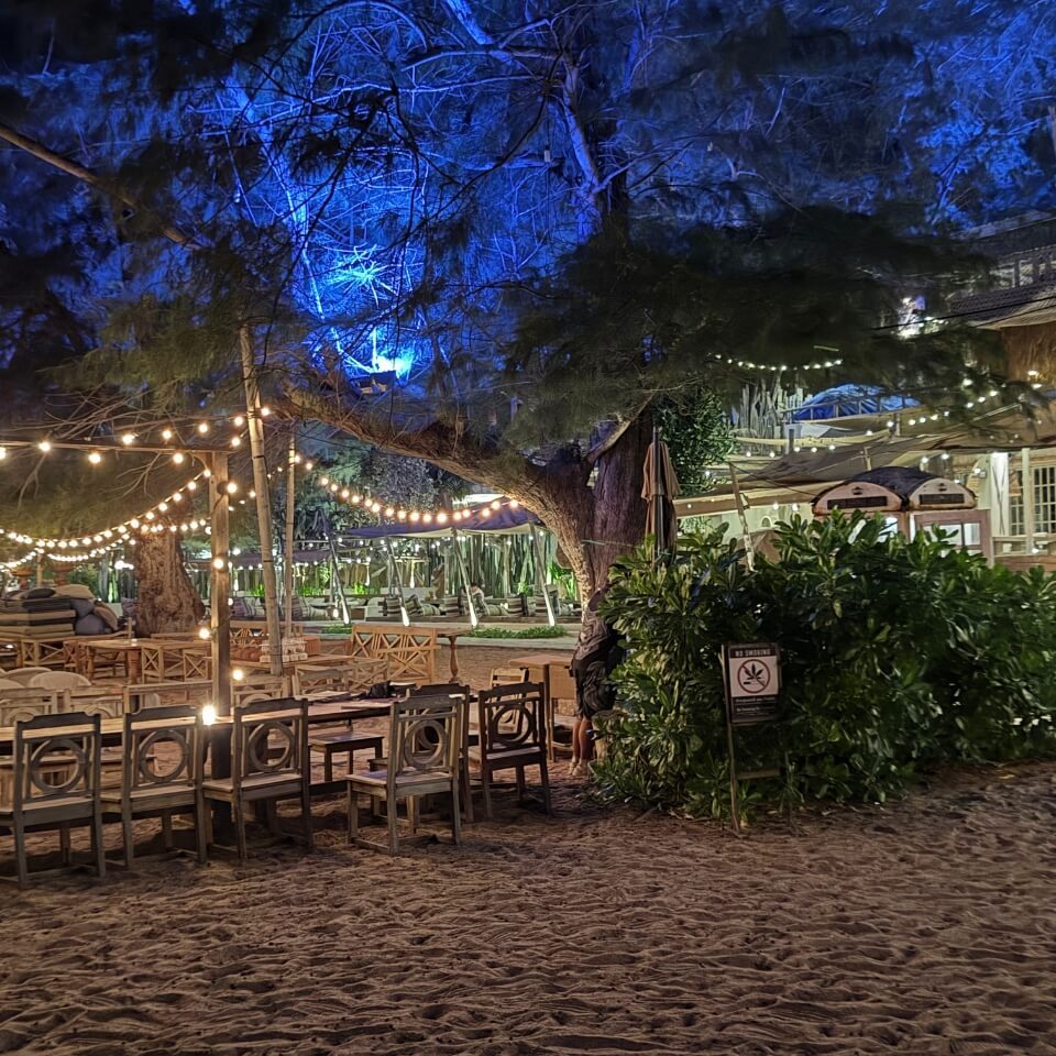 Sitzbereich am Strand mit Tischen, Stühlen, Lichterketten und Bäumen bei Nacht