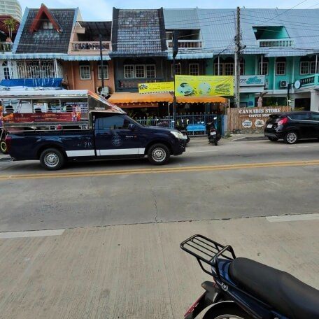 Blauer Baht Bus auf einer Straße in Jomtien mit Häusern, Geschäften und geparktem Roller im Vordergrund