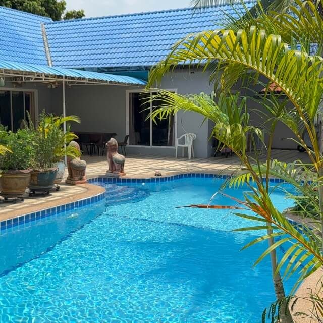Poolbereich einer Villa mit blauem Swimmingpool, Palmen, Sitzbereich und Gebäude im Hintergrund