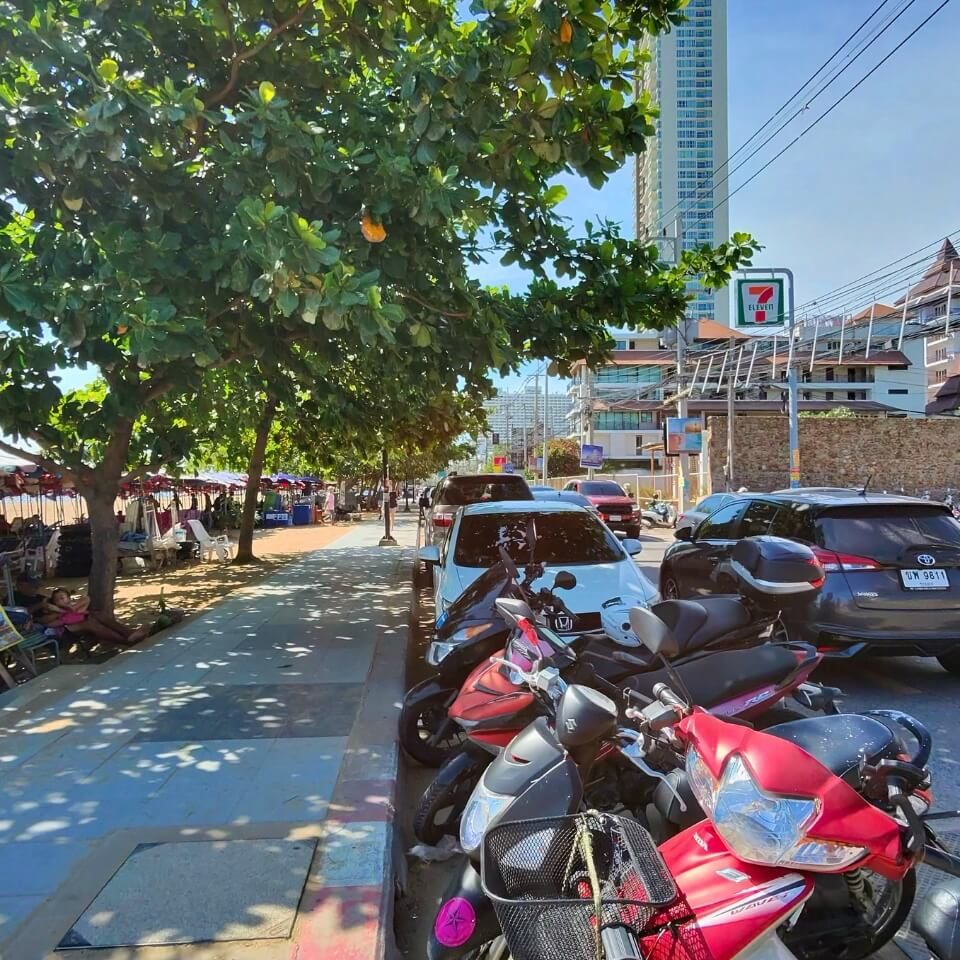 Jomtien Beach bei Tag Straße in Jomtien mit geparkten Motorrollern, schattigem Gehweg und Blick Richtung Strand bei Tageslicht