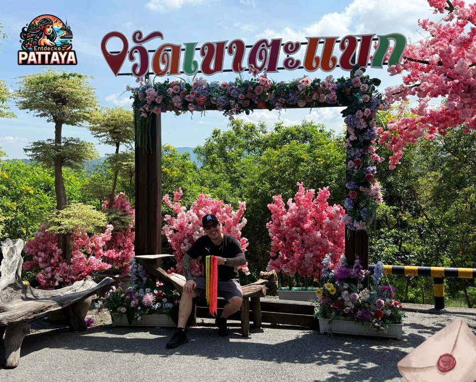 Die Pattaya Wochenmail erreicht dich auch am Skywalk Blumengeschmücktes Schild am Wat Khao Tabaek Skywalk in Si Racha mit grüner Hügellandschaft, Dennis auf einer Bank und Briefsymbol