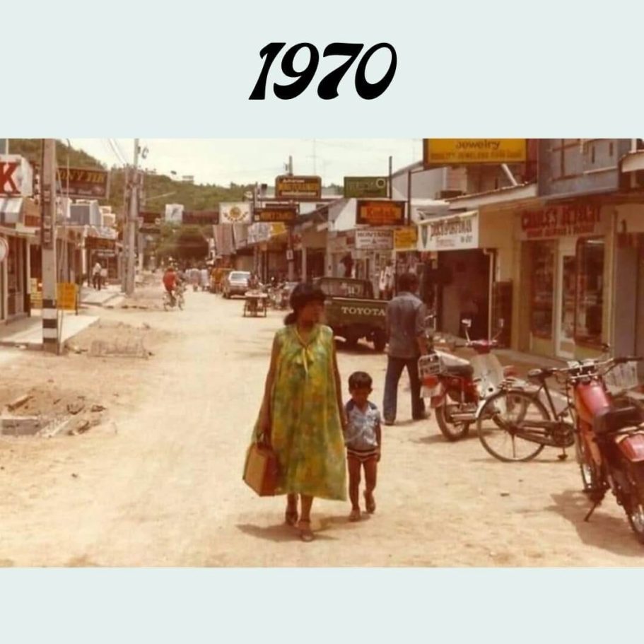 Walking Street Pattaya 1970: Eine Frau mit Kind auf der ungepflasterten Straße mit ersten Geschäften und Motorrädern