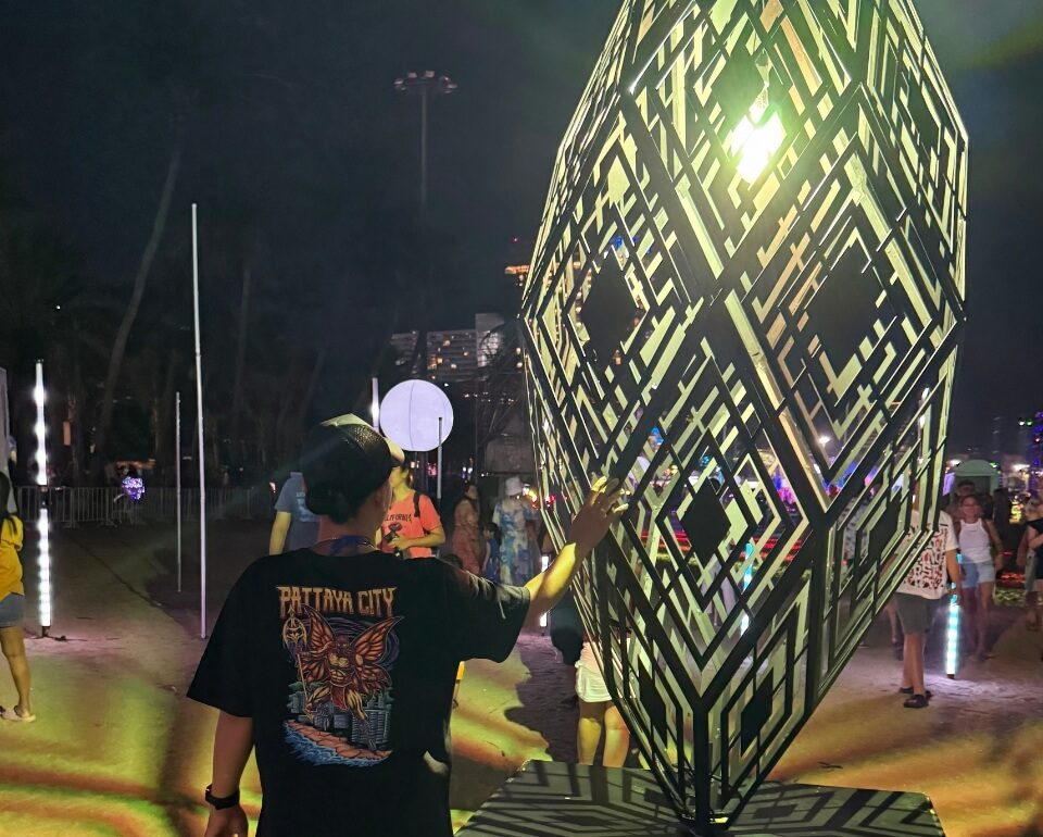 Unvergessliche Pattaya Momente für deine nächste Reise Leuchtende geometrische Skulptur bei Nacht in Pattaya mit einer Frau im Butterfly Man T-Shirt davor und weiteren Besuchern im Hintergrund