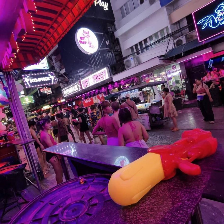Songkran 2025 auf der Soi 6 in Pattaya zene auf der Soi 6 in Pattaya während Songkran 2025 – Menschen in Badekleidung, grelle Neonlichter, eine Wasserpistole im Vordergrund.