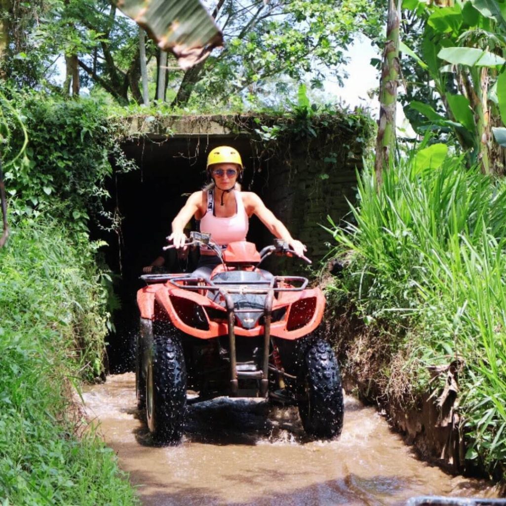Frau auf einem roten ATV fährt durch einen kleinen Wasserkanal, umgeben von üppiger grüner Natur in Thailand.